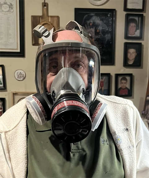 Harry Urban wearing a full-face protective respirator with filters for woodworking, seated indoors with framed photographs behind him.